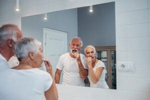 An older man and woman brushing their tooth together in front of the bathroom mirror.